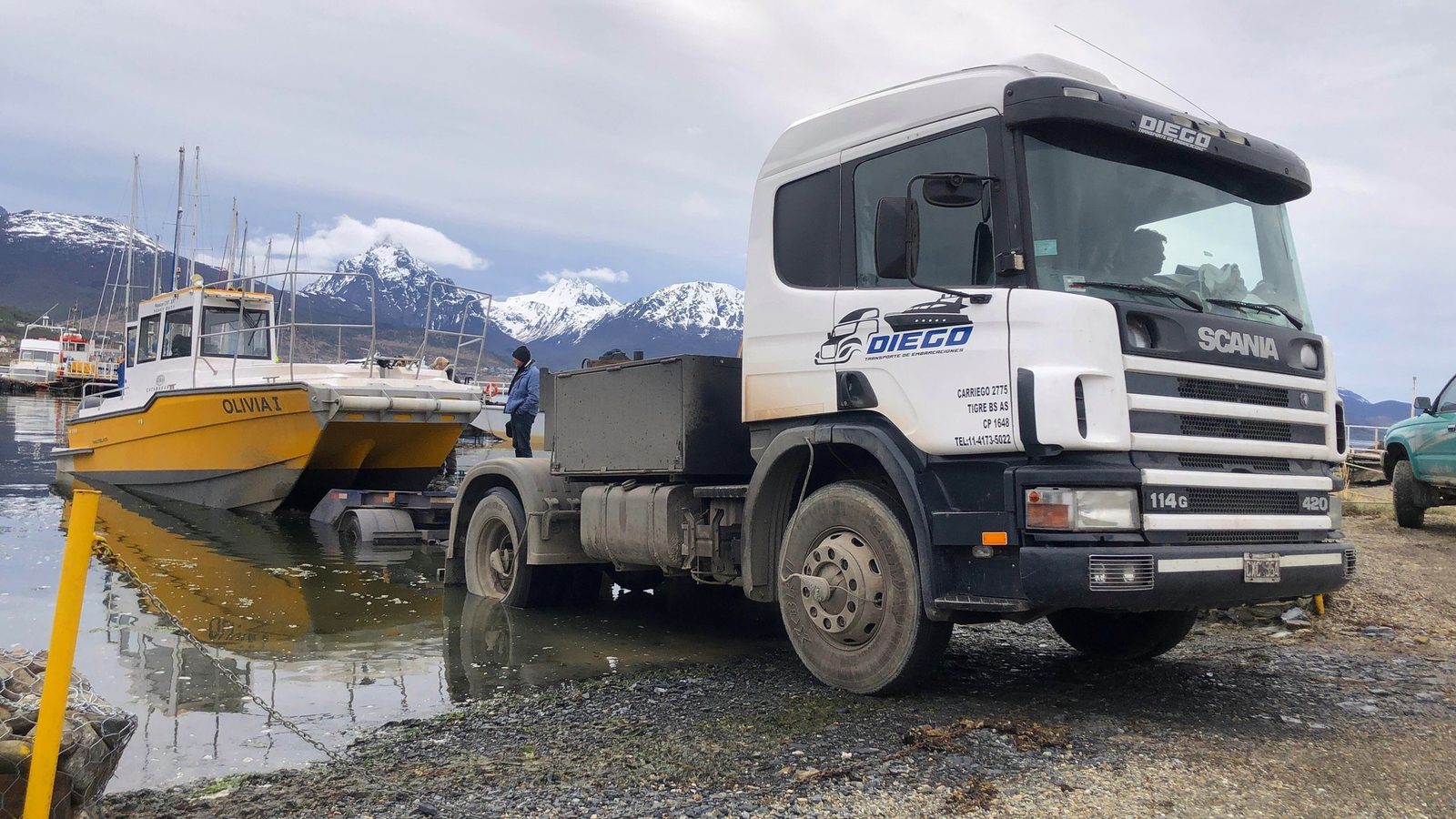 Camión Scania de Transporte Diego en la costa de Ushuaia, con una lancha amarilla y la cordillera nevada
